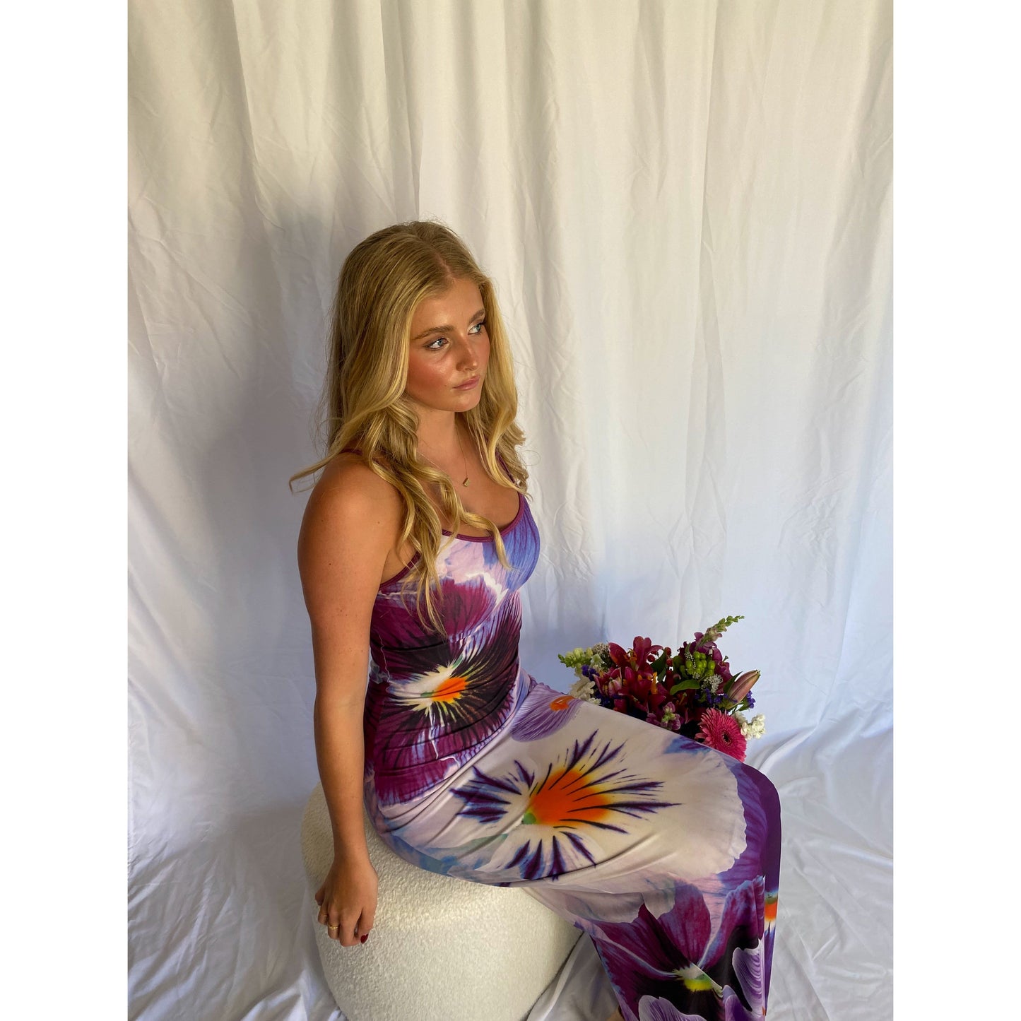 With Harper Lu Indigo Slip, Size S. Featuring an accentuated waistline, adjustable straps, a rear split, and fully lined, this dress is designed to skim the body. With an amethyst print with a few pops of tangelo. Perfect for formals, birthdays, etc. Front on sitting view of dress.